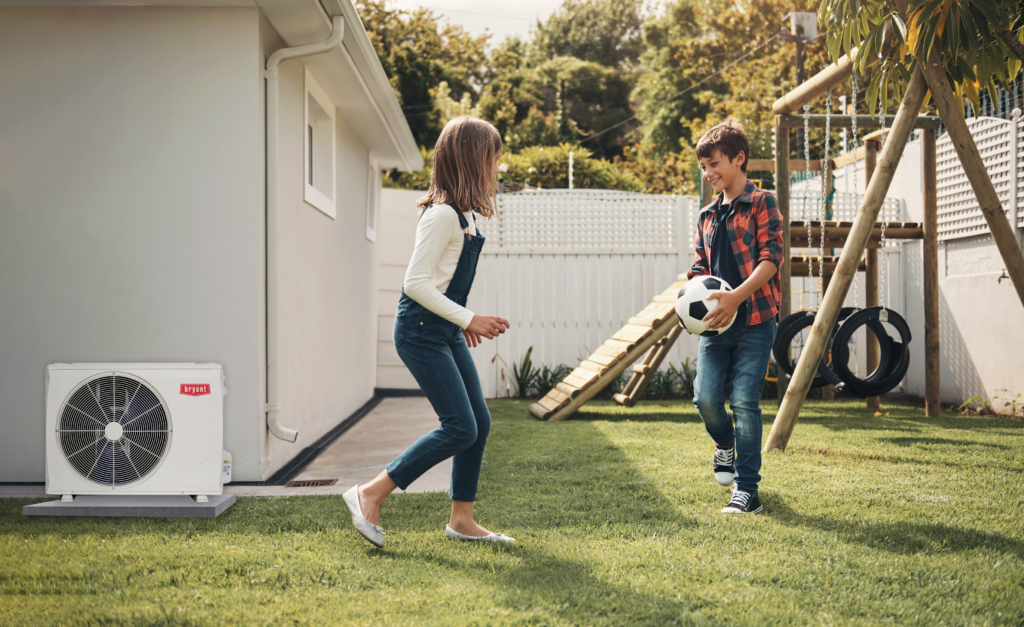 Kids happily playing with a soccer ball outside in their yard with a Bryant air conditioner nearby, representing the benefits of a new HVAC system.
