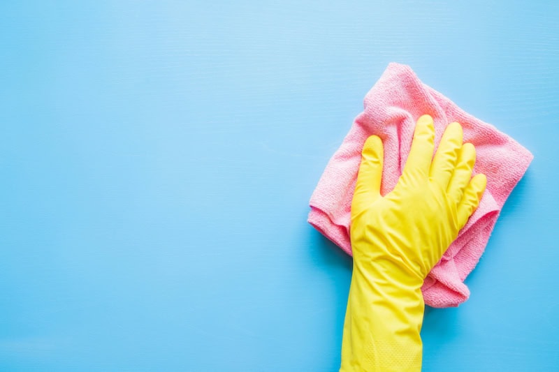 Photo of a hand in a yellow rubber glove, wiping a blue wall or table with a pink cloth.