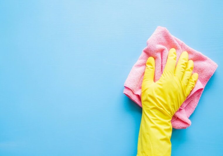 Photo of a hand in a yellow rubber glove, wiping a blue wall or table with a pink cloth.